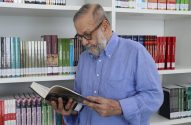 Homem idoso de óculos e barba lendo um livro em uma biblioteca.