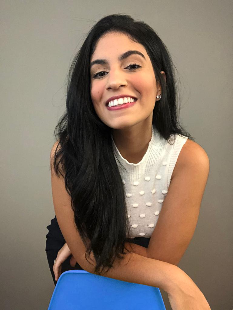 Mulher jovem sorridente com braços apoiados em cadeira azul. Veste blusa branca. Fundo cinza.