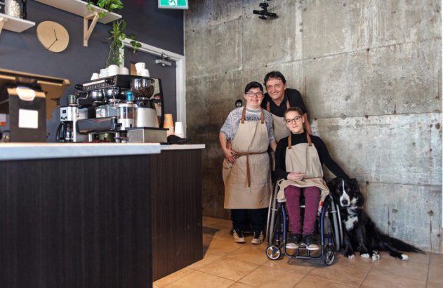 Três baristas (jovem com Síndrome de Down, jovem em cadeira de rodas, homem) e um cão em cafeteria.