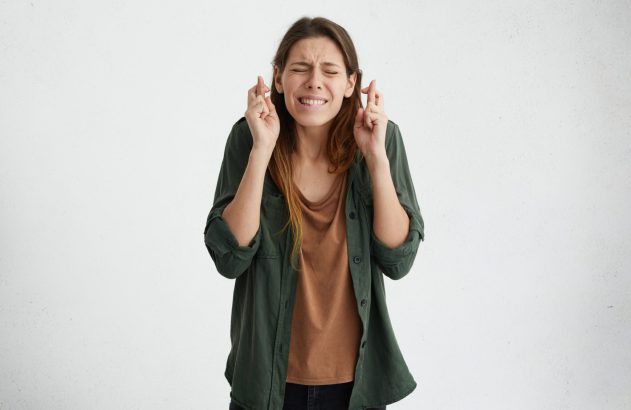 Mulher de olhos fechados e feição contraída, fazendo um pedido com os dedos cruzados.
