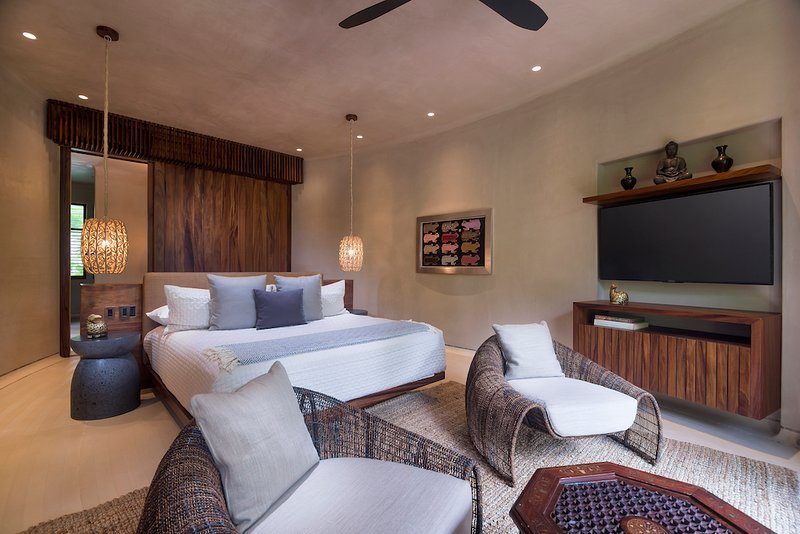 Quarto com cama e cabeceira de madeira, TV na parede, poltronas de vime e luminárias pendentes.