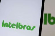Close-up de smartphone exibindo logo 'Intelbras' verde na tela branca. Reflexo e mão segurando aparelho.
