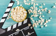 Uma tigela amarela de pipoca e uma claquete de cinema sobre uma superfície de madeira azul-turquesa, com pipoca espalhada.