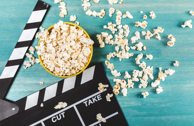 Uma tigela amarela de pipoca e uma claquete de cinema sobre uma superfície de madeira azul-turquesa, com pipoca espalhada.