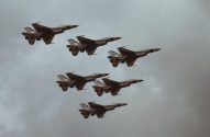 Sete jatos F-16 em formação no céu nublado. Aeronaves com pintura escura e clara, detalhes de flor de cerejeira.