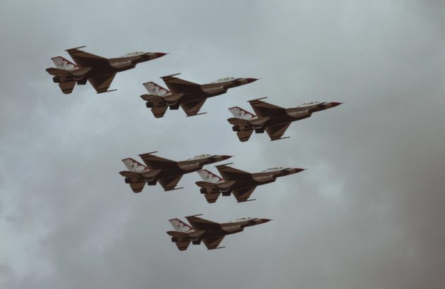 Sete jatos F-16 em formação no céu nublado. Aeronaves com pintura escura e clara, detalhes de flor de cerejeira.