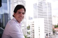Homem sorrindo, olhando para a câmera, com prédios da cidade desfocados ao fundo.
