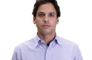 Homem de frente, cabelo escuro e camisa roxa clara, em fundo branco.