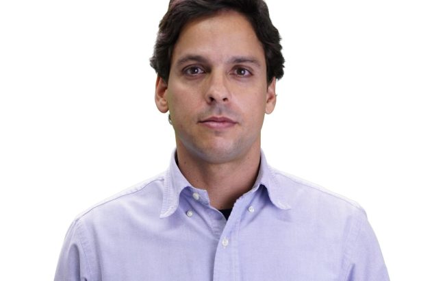 Homem de frente, cabelo escuro e camisa roxa clara, em fundo branco.