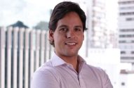 Homem sorrindo em camisa xadrez, com prédios urbanos desfocados ao fundo.