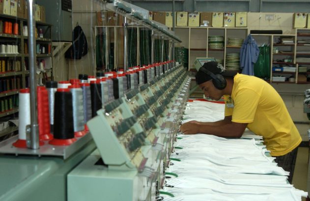 Homem opera máquina de bordar industrial em fábrica. Camisas brancas recebem bordados coloridos. Fios e fones de ouvido visíveis.