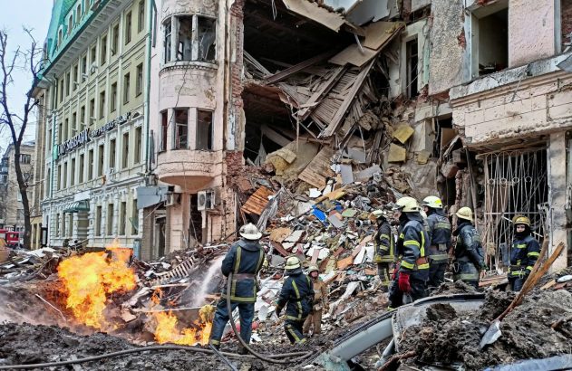 Bombeiros combatem fogo entre escombros de edifício destruído. Área urbana em ruínas, com fumaça.