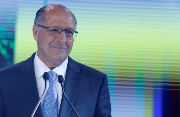 Homem de terno azul e óculos sorrindo em pódio com microfones. Fundo desfocado com faixas de luz verde e azul.