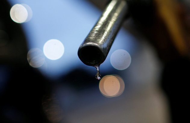 Uma gota de combustível pingando de um bico de bomba, com luzes bokeh desfocadas ao fundo.