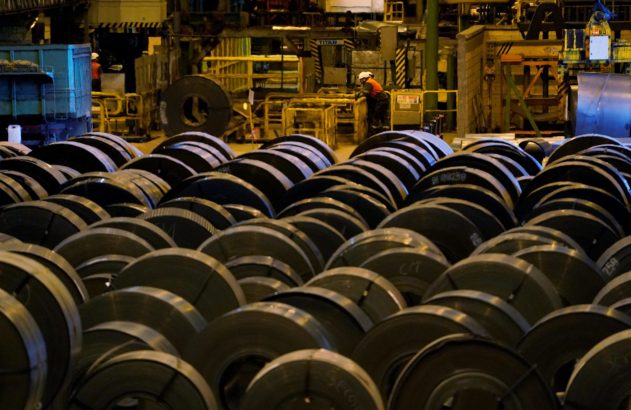 Bobinas de aço empilhadas em chão de fábrica com operários e máquinas ao fundo.