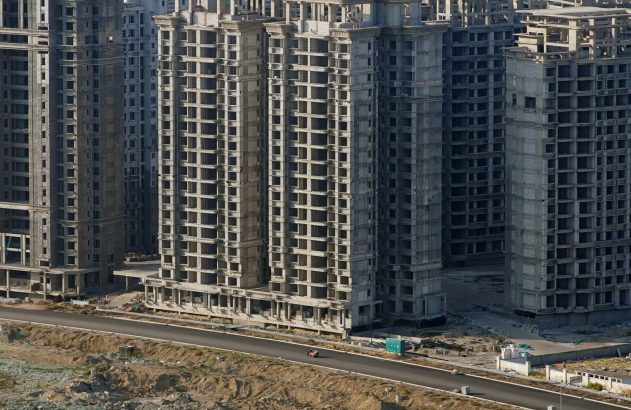Vista aérea: vários prédios de concreto inacabados. Uma moto em estrada deserta entre a construção.