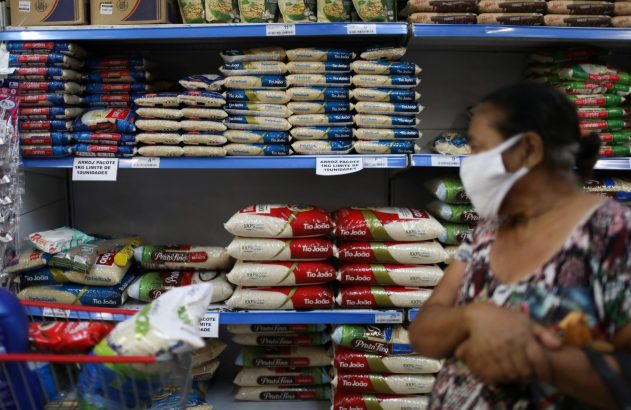 Mulher de máscara compra arroz em supermercado, onde prateleiras cheias exibem limite de 10 pacotes.