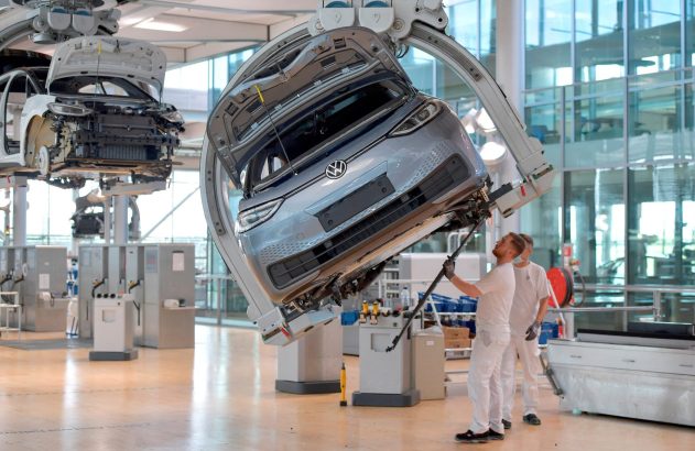 Carro Volkswagen cinza é suspenso por braço robótico em fábrica; operários trabalham na parte inferior do veículo.