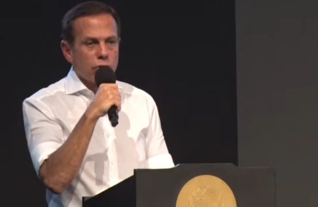 Homem de camisa branca discursa ao microfone, atrás de púlpito com emblema dourado.