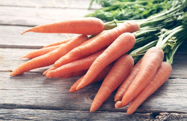 Cenouras frescas e laranjas com talos verdes, dispostas sobre uma mesa de madeira rústica.