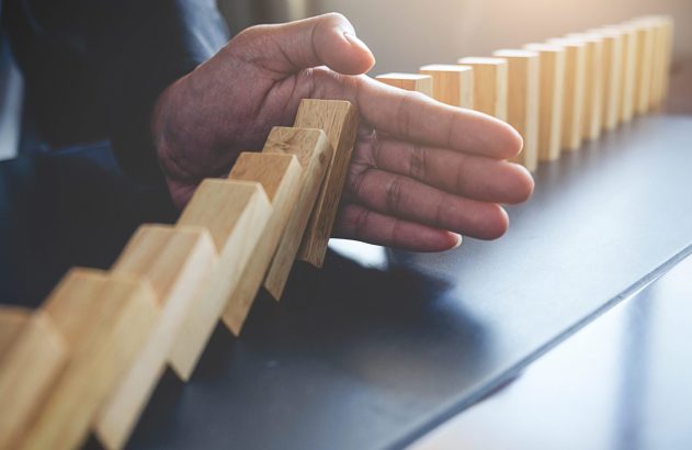 Mão de pessoa impedindo a queda de dominós de madeira, parando a reação em cadeia.