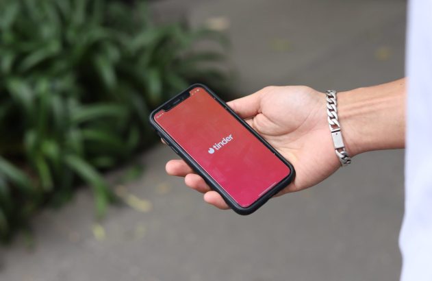 Mão segura smartphone exibindo o logo e nome do app Tinder em tela cheia. Pulso com pulseira.