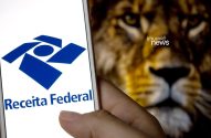 Mão segura smartphone com logo da Receita Federal. Fundo desfocado mostra rosto de leão.