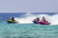 Três jet skis com pilotos na água, gerando grande spray ao acelerar.