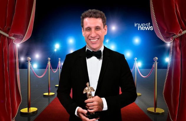 Homem de smoking sorridente segura troféu em tapete vermelho, com cortinas e luzes. Logo Invest News no canto.