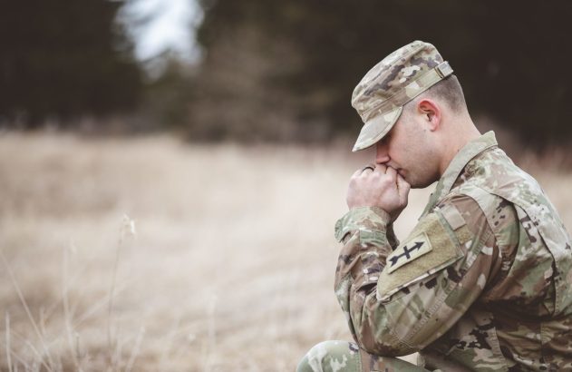 Militar uniformizado, cabisbaixo com mãos unidas, ajoelhado em campo.