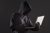 Pessoa encapuzada com luvas pretas, usando um laptop branco e digitando. O rosto está oculto pelo capuz, em fundo escuro.