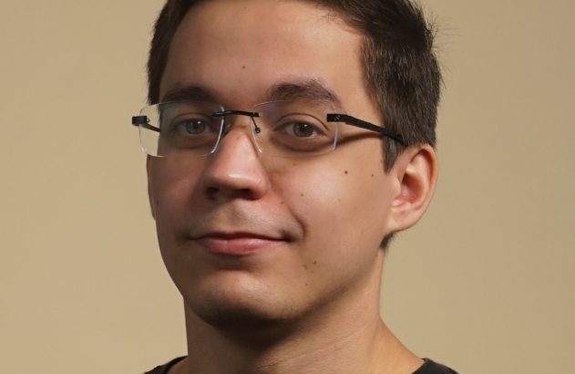 Retrato de homem jovem com óculos, camiseta preta e leve sorriso.