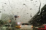 Trânsito em dia chuvoso visto por parabrisa com gotas d'água e luzes de freio de carros.