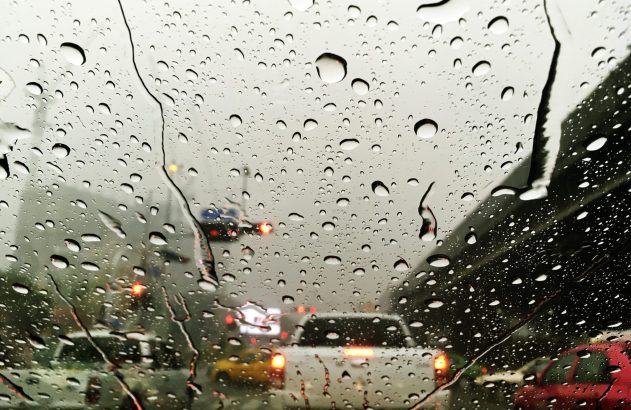 Trânsito em dia chuvoso visto por parabrisa com gotas d'água e luzes de freio de carros.