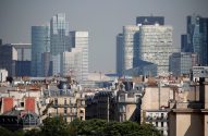 Telhados parisienses em primeiro plano, com arranha-céus da La Défense ao fundo sob céu claro.