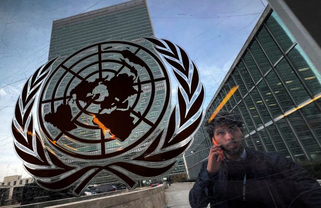 Emblema da ONU e edifício da Sede da ONU; reflexo de homem no telefone em vidro riscado.