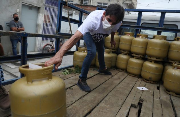 Homem mascarado tira papel de botijão de gás amarelo em caminhão, com pilhas de botijões.