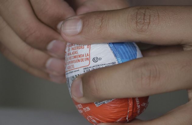 Mãos segurando um ovo de chocolate. A embalagem colorida exibe um aviso de segurança sobre supervisão adulta.