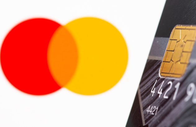 Cartão de crédito escuro com chip dourado e números visíveis em primeiro plano, com o logo desfocado da Mastercard ao fundo.