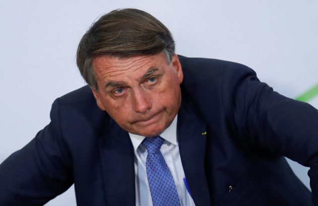 Jair Bolsonaro, de terno azul-marinho e gravata, com expressão séria, inclina-se levemente.