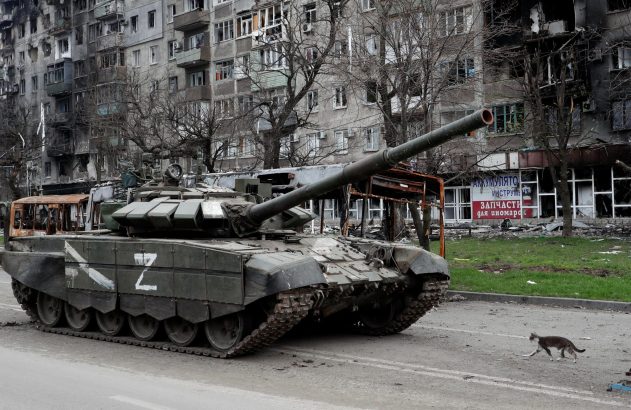 Um tanque com marca 'Z' em rua de cidade destruída. Um gato cruza a via.