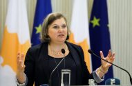 Victoria Nuland gesticula com as mãos abertas ao falar em púlpito. Bandeiras de Chipre e UE ao fundo.