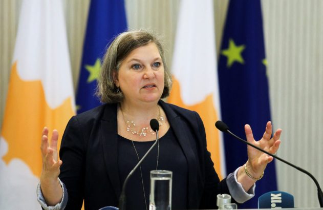 Victoria Nuland gesticula com as mãos abertas ao falar em púlpito. Bandeiras de Chipre e UE ao fundo.