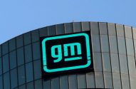 Logotipo neon verde da GM em prédio moderno de vidro sob céu azul.
