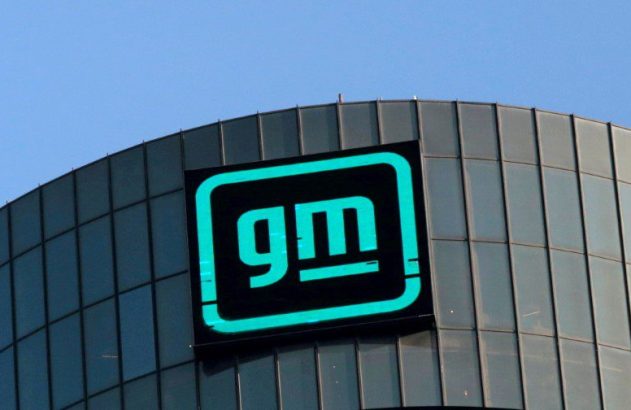 Logotipo neon verde da GM em prédio moderno de vidro sob céu azul.