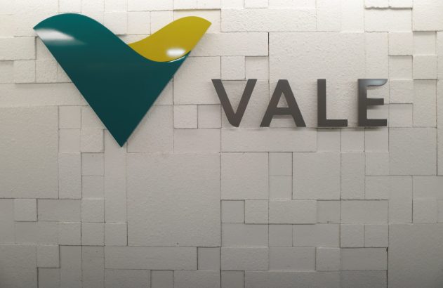 Logotipo da VALE em verde e amarelo e nome "VALE" em cinza, sobre uma parede branca texturizada com blocos.