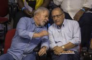 Lula (camisa jeans) e Alckmin (óculos) conversam sentados. Lula gesticula, Alckmin ouve.