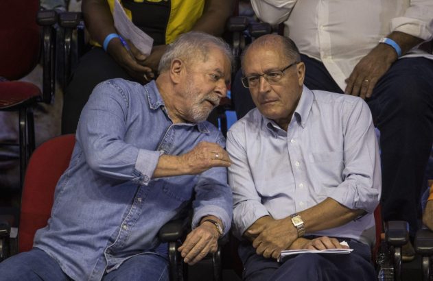 Lula (camisa jeans) e Alckmin (óculos) conversam sentados. Lula gesticula, Alckmin ouve.