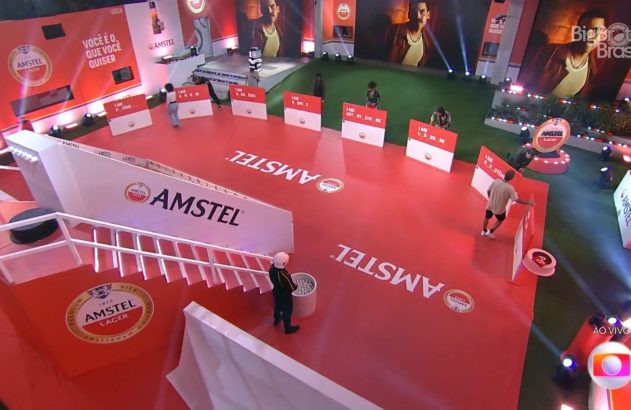 Participantes em desafio Amstel no Big Brother Brasil. Cenário vermelho com logos e placas "I AM...", e escadaria branca.