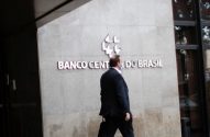 Homem de terno caminha em frente ao Banco Central do Brasil, com chão molhado e reflexivo.